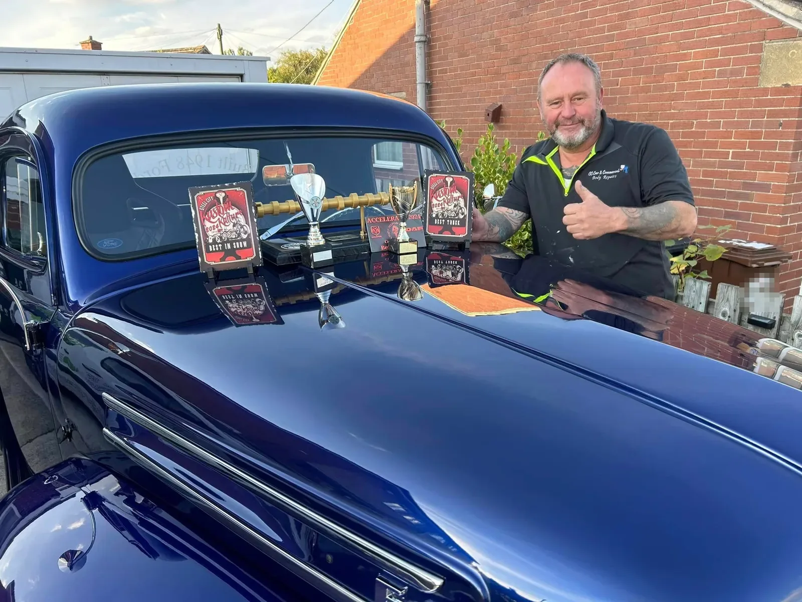 A man in a CC Car & Commercial polo shirt giving a thumbs up beside a deep blue Ford F100, with Best in Show and Best Truck trophies on the bonnet