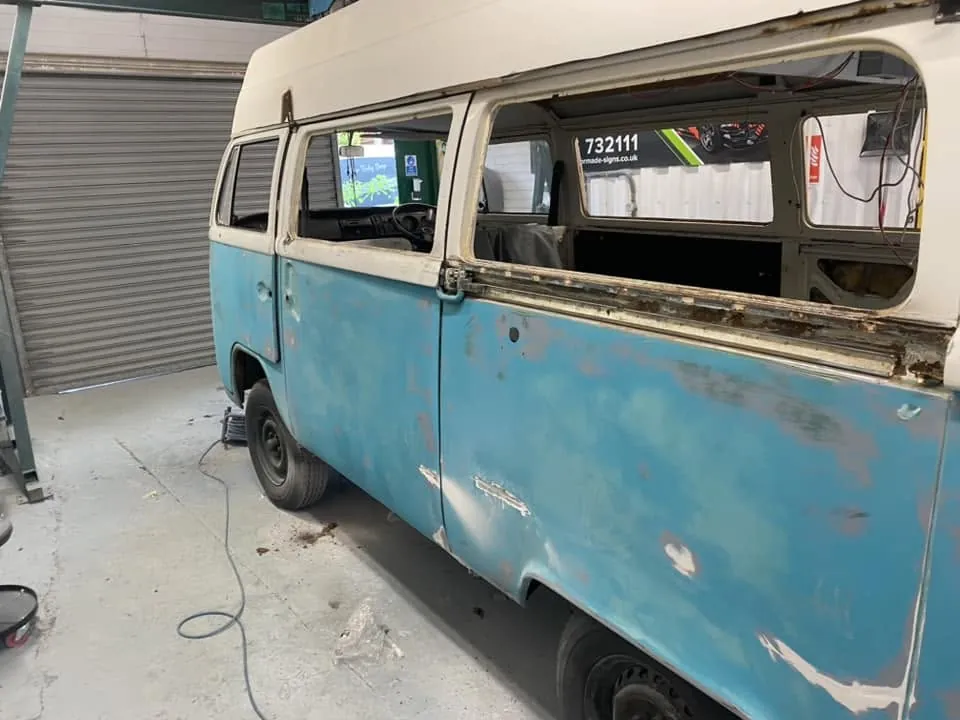 A turquoise and white VW camper in the workshop, paint flaking and rust along its side